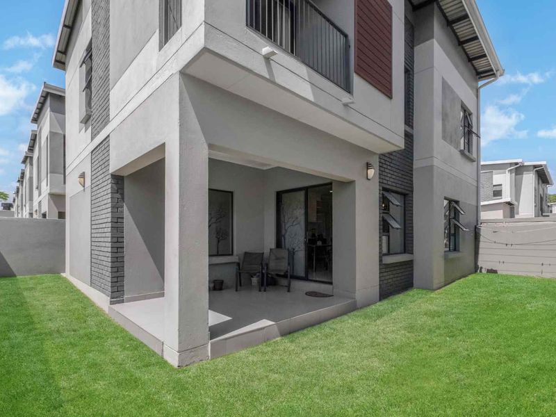 A modern rear exterior view of a cluster home featuring a covered patio, tiled flooring, and a private garden area.