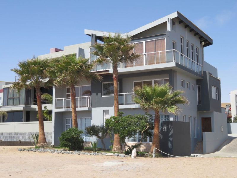 A modern multi-story residential building featuring balconies with glass balustrades, large sliding doors, and contemporary architectural design, situated near a sandy beach area.