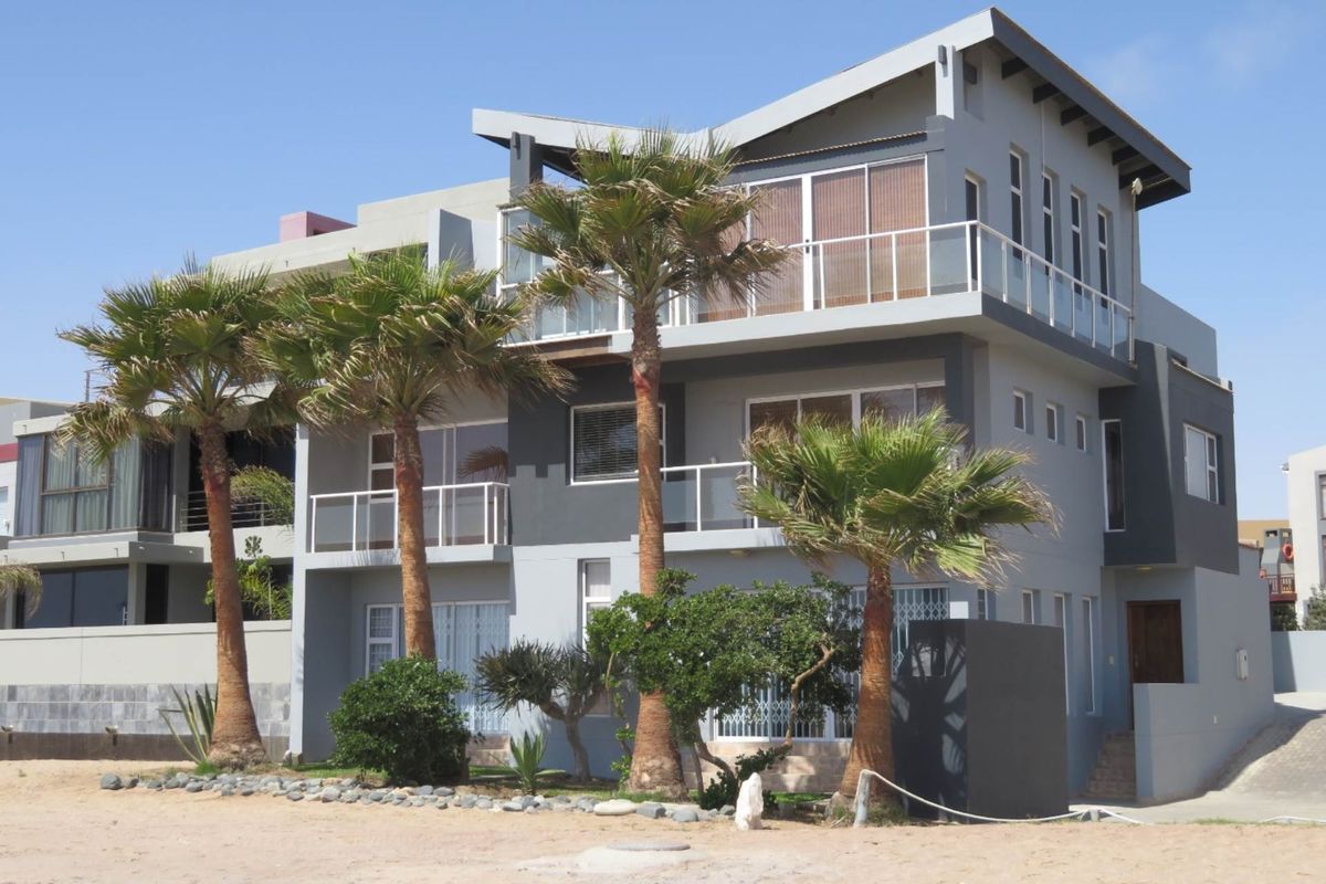 A modern multi-story residential building featuring balconies with glass balustrades, large sliding doors, and contemporary architectural design, situated near a sandy beach area.