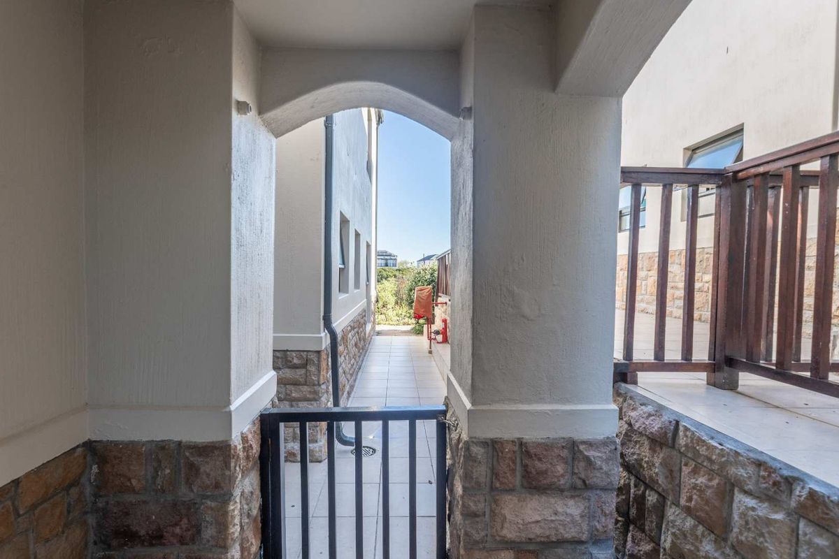 A view of a paved exterior walkway featuring stone-clad pillars, a metal security gate, and a timber balustrade leading to a rear outdoor area.