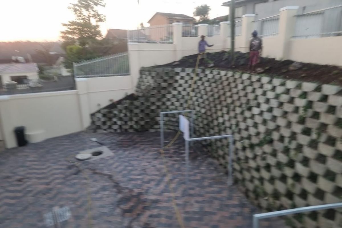 Rear exterior area featuring a large tiered retaining wall, paved ground surface, and perimeter boundary wall with metal fencing.