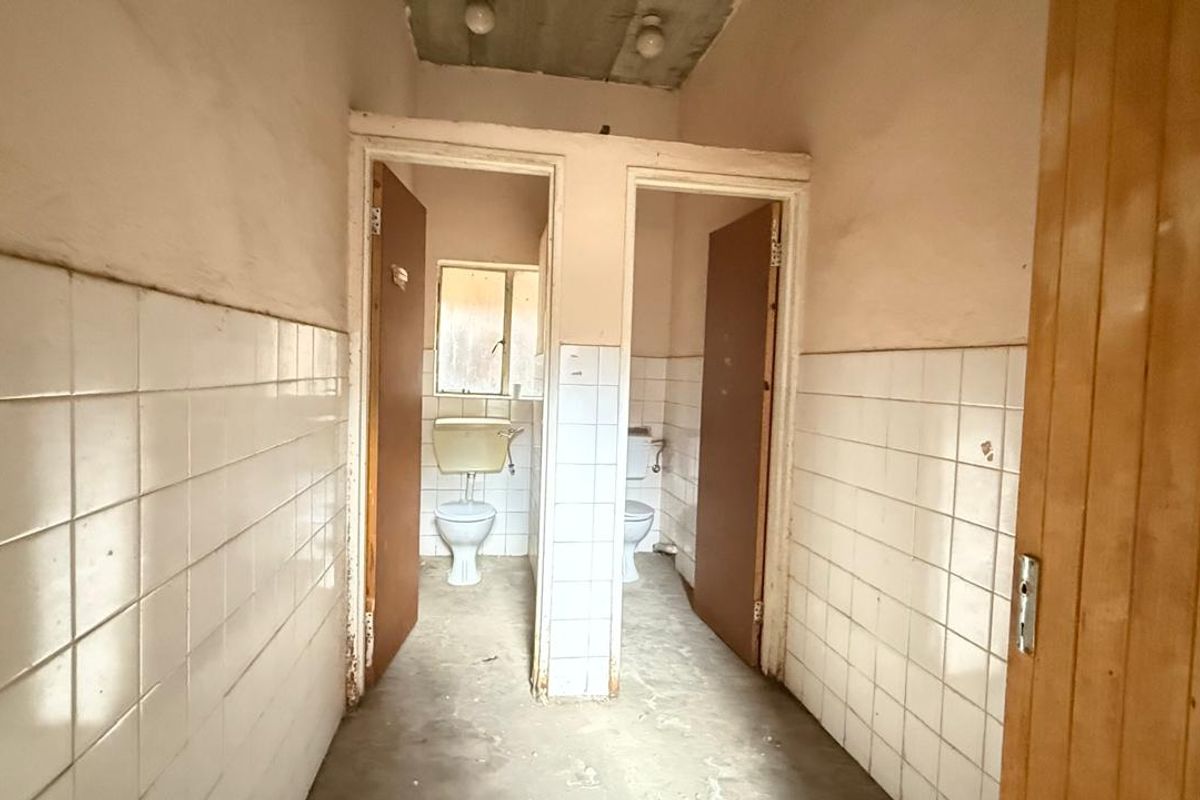 A dated bathroom facility featuring two separate toilet cubicles with tiled walls and unfinished ceiling, requiring significant renovation and modernization.