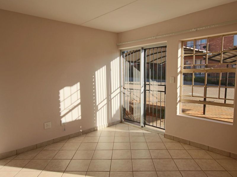 An empty living area featuring neutral tiled flooring, a sliding glass door with security gate, and natural light through a window with security bars.