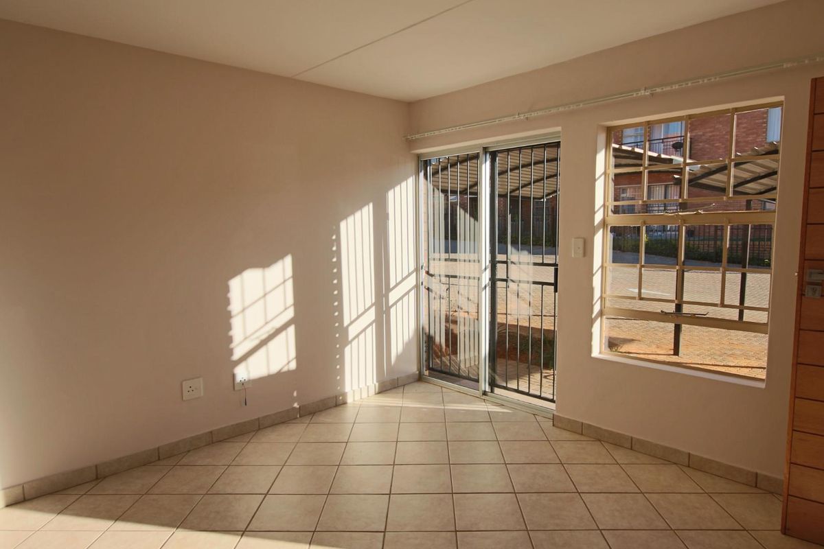 An empty living area featuring neutral tiled flooring, a sliding glass door with security gate, and natural light through a window with security bars.