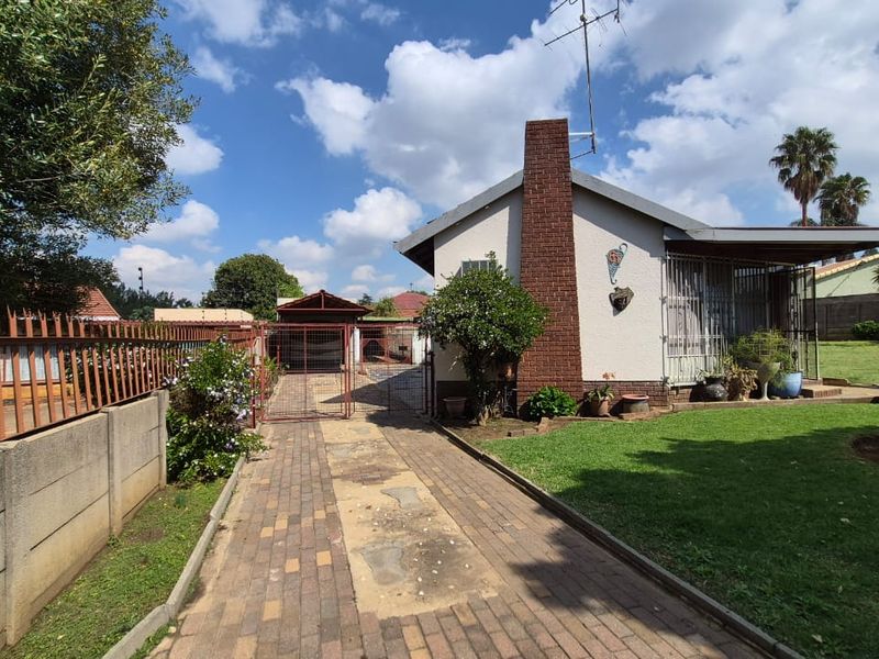 Front view of the property featuring a paved driveway, brick chimney, covered patio area, and established garden with perimeter wall and metal fencing.
