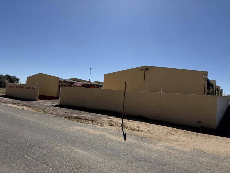 Street view of the apartment complex entrance featuring a perimeter wall with electric fencing and a paved driveway area.