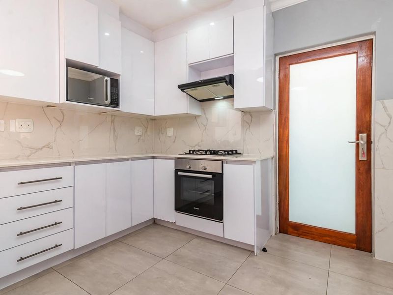 Modern kitchen featuring white high-gloss cabinetry, marble-look backsplash, integrated gas hob, oven, extractor fan, and a frosted glass door leading outside.