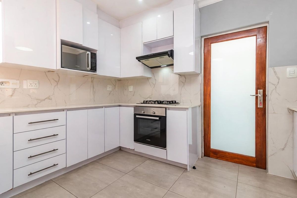 Modern kitchen featuring white high-gloss cabinetry, marble-look backsplash, integrated gas hob, oven, extractor fan, and a frosted glass door leading outside.