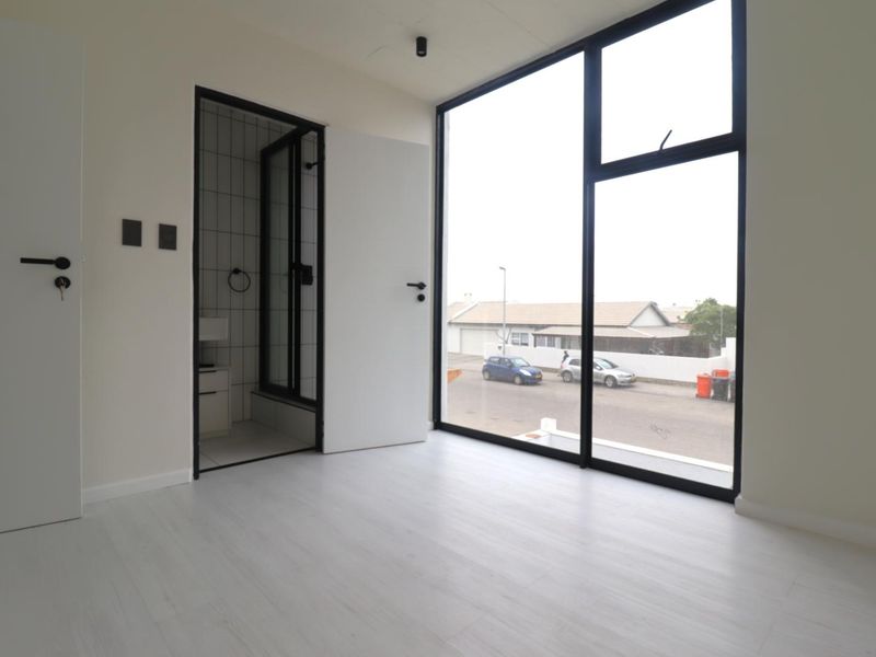 A bright, modern bedroom featuring large floor-to-ceiling windows, light laminate flooring, and direct access to an en-suite bathroom.