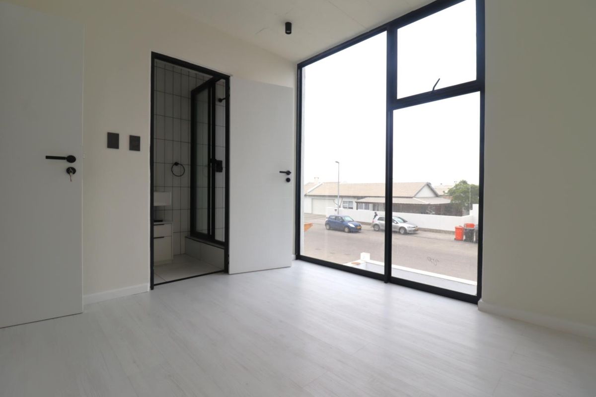 A bright, modern bedroom featuring large floor-to-ceiling windows, light laminate flooring, and direct access to an en-suite bathroom.