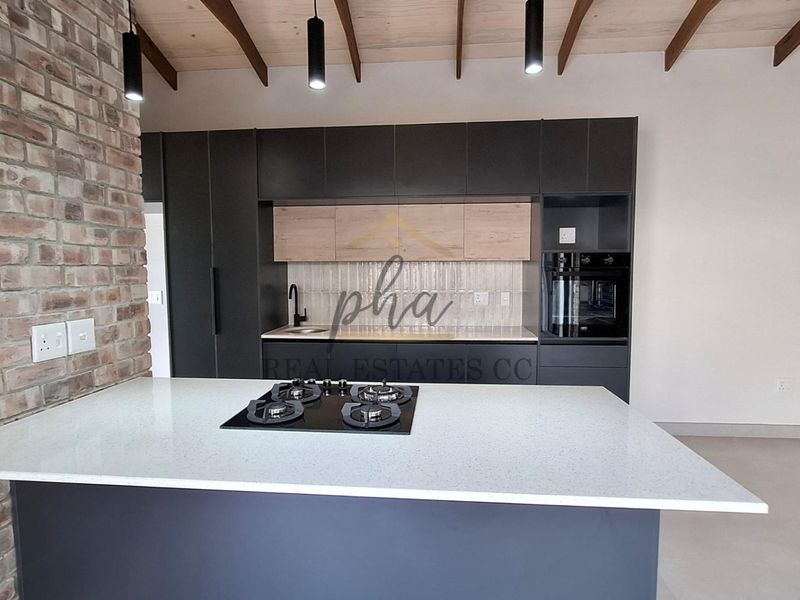 Modern kitchen featuring dark cabinetry, stone countertops, a central island with a gas hob, exposed brickwork, and timber ceiling beams.