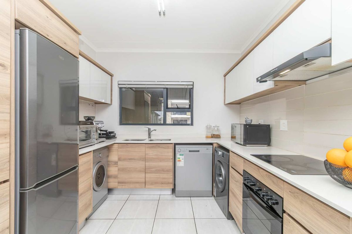 Modern galley-style kitchen featuring light wood-tone cabinetry, white stone countertops, tiled backsplash, and integrated appliances including an electric hob and oven.