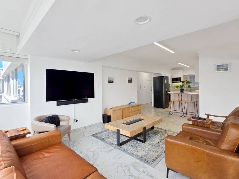 A bright open-plan living and kitchen area featuring modern marble-look floor tiles, recessed lighting, and a functional kitchen island with built-in cabinetry.
