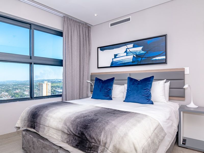 Modern bedroom featuring large windows with expansive city views, ducted air conditioning, recessed lighting, and neutral tiled flooring.