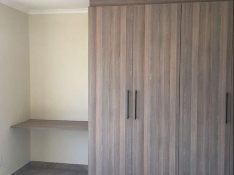 A clean bedroom featuring modern wood-finish built-in wardrobes and a matching integrated desk, complemented by neutral tiled flooring.