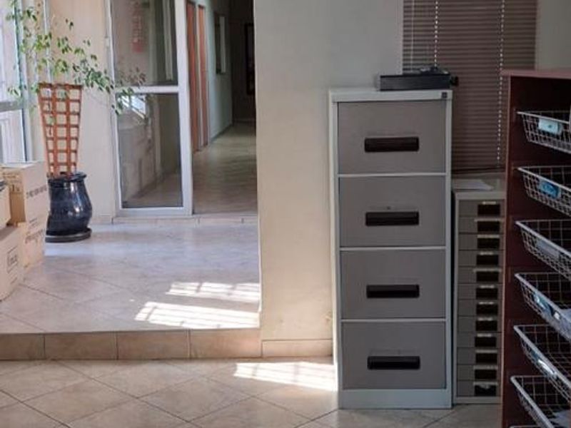 An office space featuring tiled flooring and venetian blinds, with a clear view through to an adjacent hallway area.