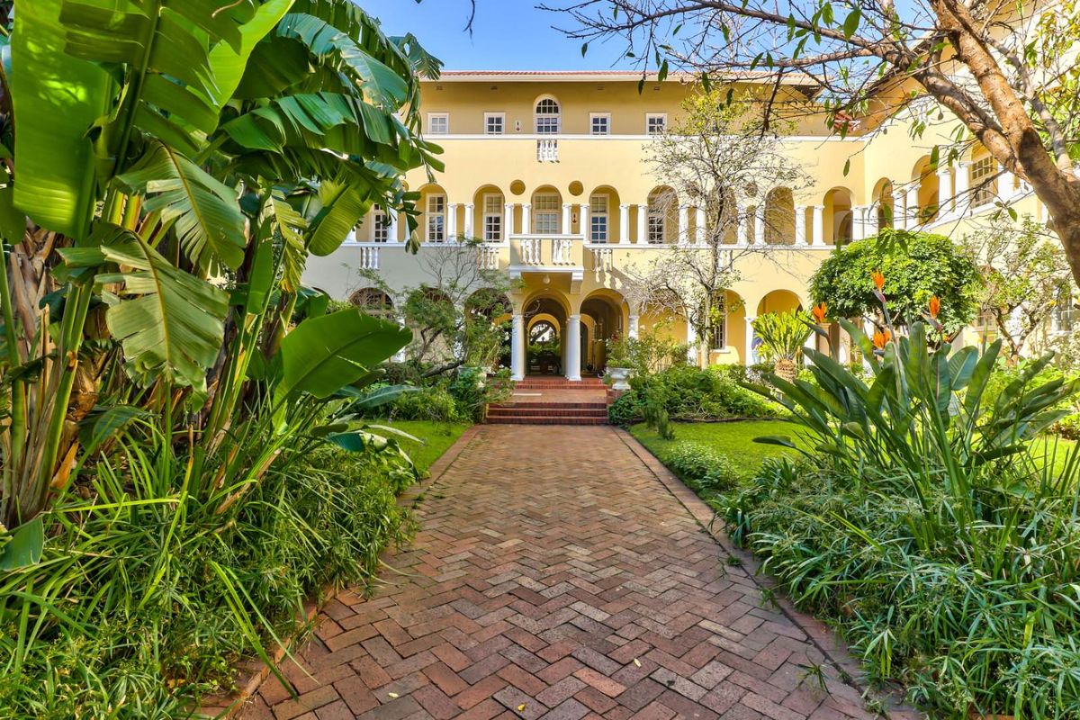 A beautifully landscaped courtyard garden featuring a brick pathway leading to a classic Mediterranean-style apartment building with arched colonnades and balconies.