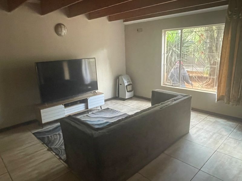 A living room featuring exposed timber ceiling beams, tiled flooring, and a large window with security burglar bars.