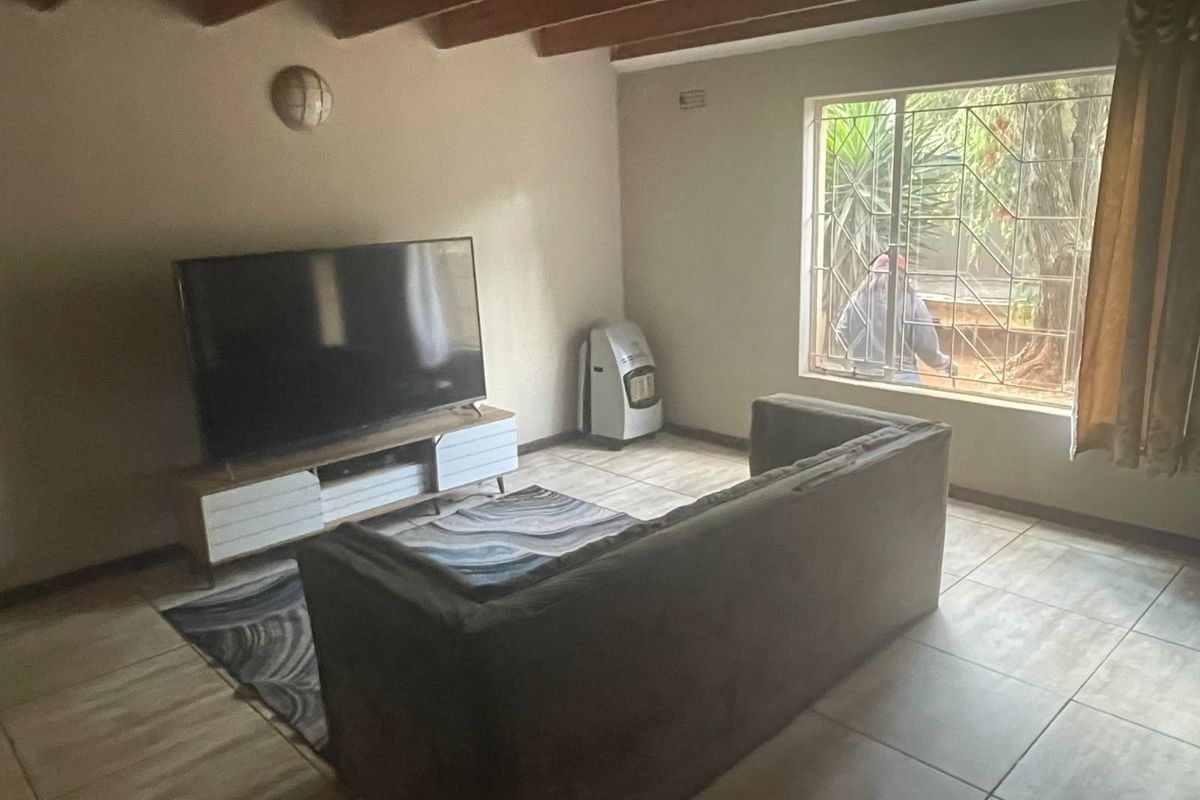 A living room featuring exposed timber ceiling beams, tiled flooring, and a large window with security burglar bars.