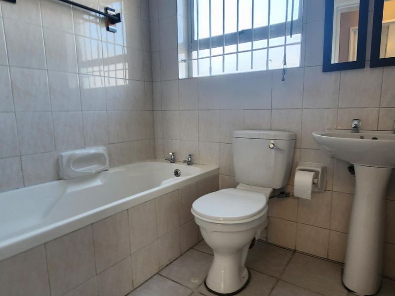 A functional bathroom featuring a standard bathtub, pedestal basin, and toilet with tiled finishes and security bars on the window.