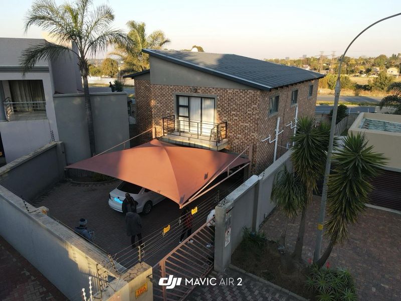 Aerial view of a two-story brick house featuring a paved driveway with a shade sail, a balcony, and secure perimeter walling with electric fencing.