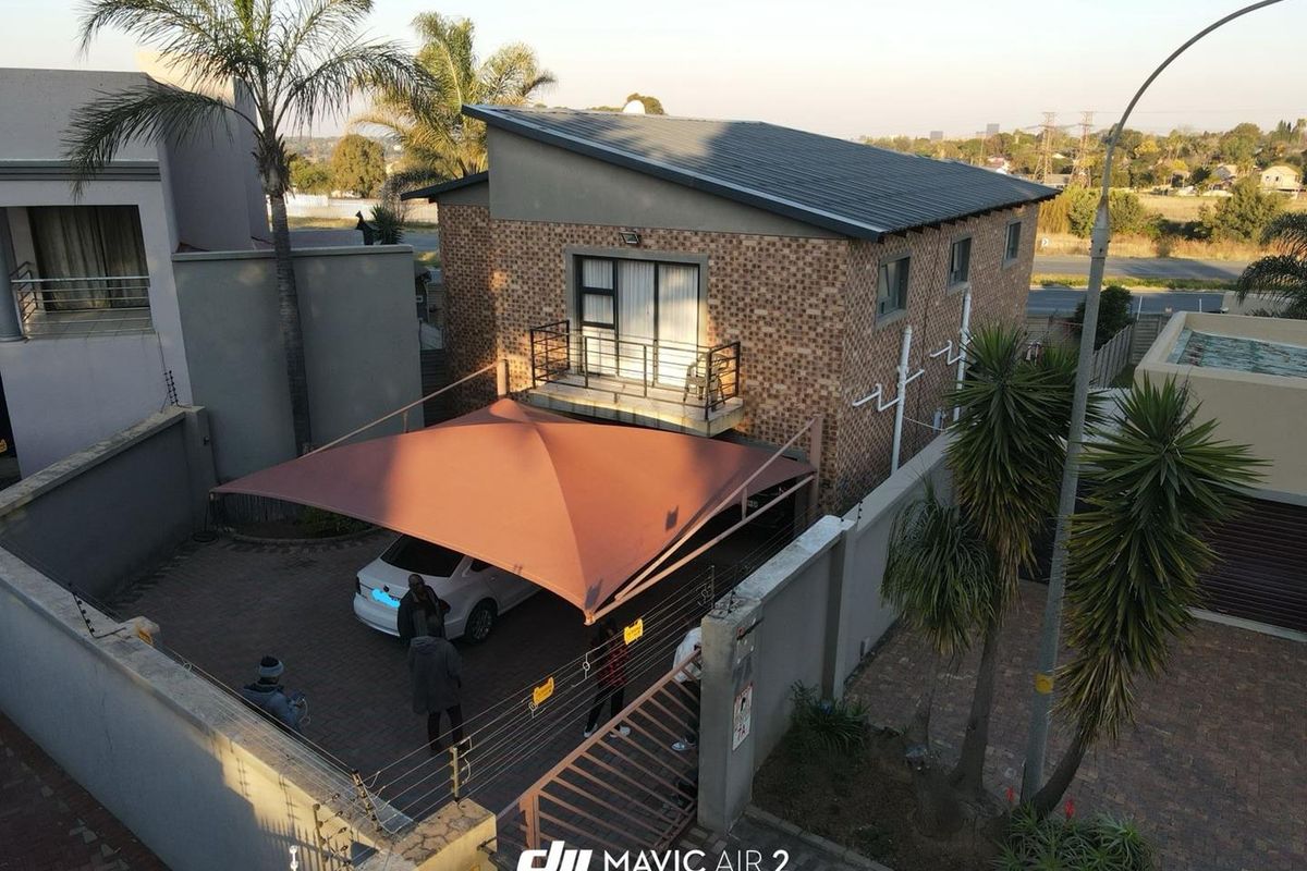 Aerial view of a two-story brick house featuring a paved driveway with a shade sail, a balcony, and secure perimeter walling with electric fencing.