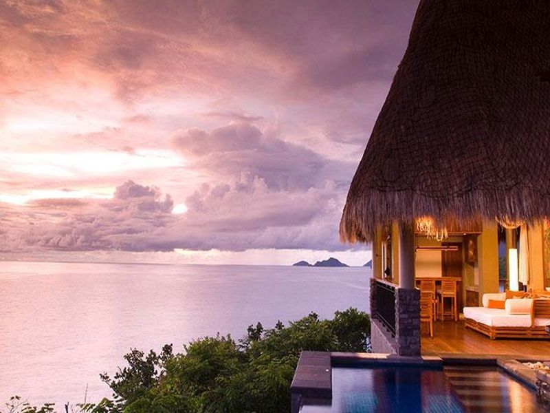 A stunning infinity pool overlooking the ocean at sunset, featuring a covered patio area with a traditional thatched roof and timber decking.