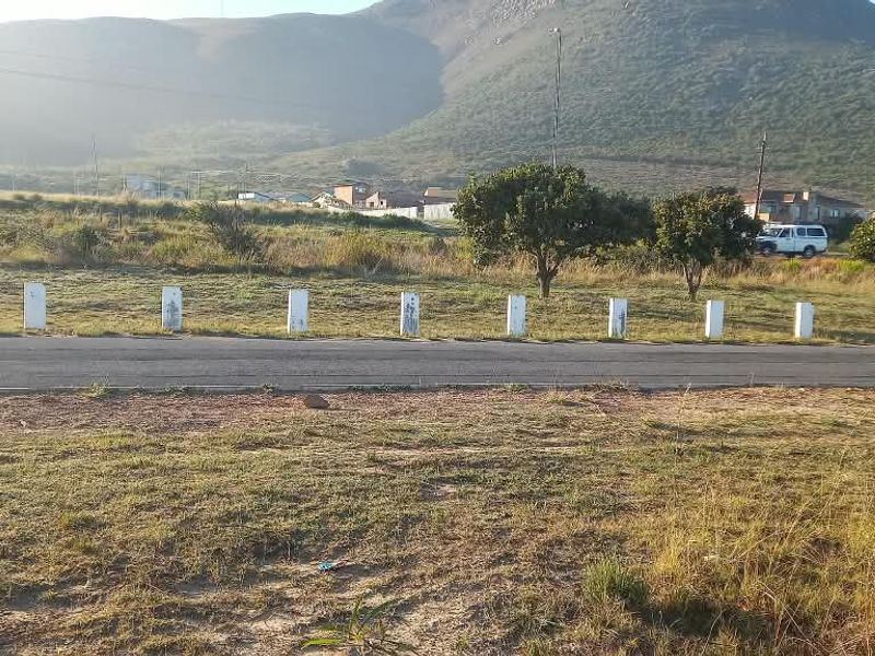 A view of a vacant residential plot featuring a paved road frontage and boundary bollards, set against a scenic mountain backdrop.