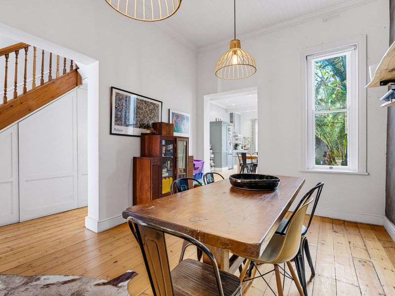 Dining room featuring polished timber flooring, a classic timber staircase, and pendant lighting, with a view through to the kitchen area.
