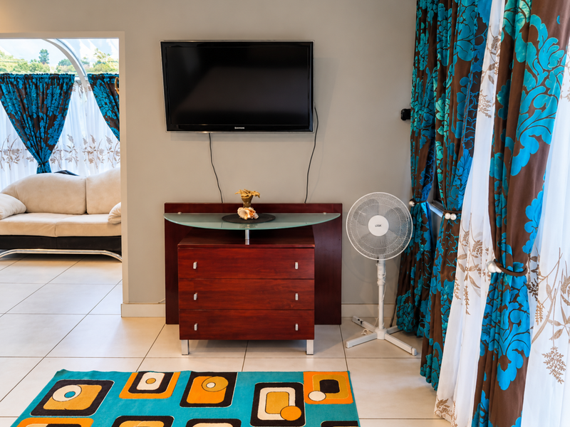 A living room area featuring tiled flooring and a wall-mounted television, with an adjacent view into a secondary lounge space.