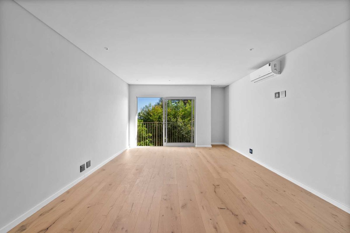A bright, empty living room featuring light timber flooring, recessed lighting, a split-system air conditioner, and a sliding glass door leading to a balcony.