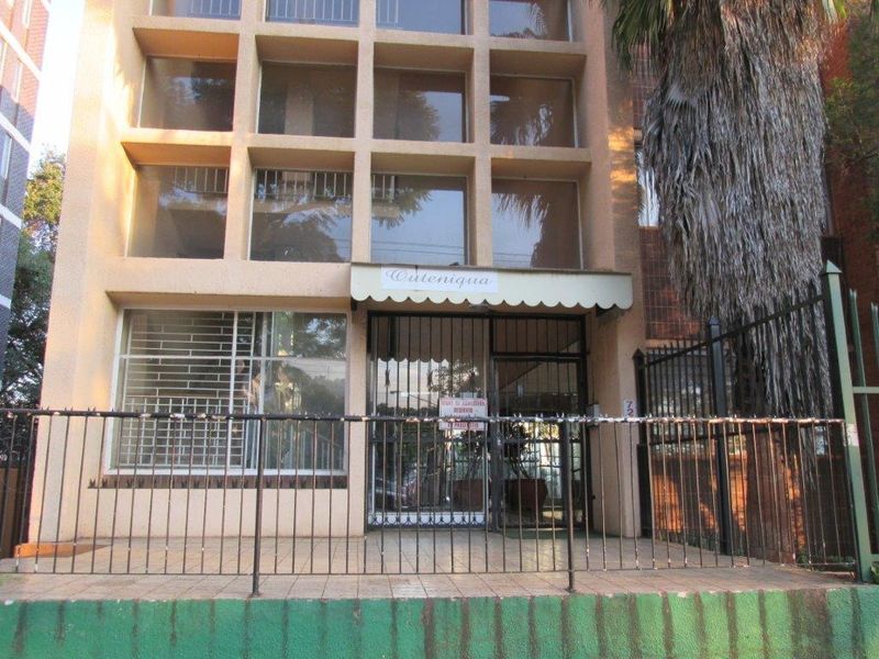 Front facade of the apartment building featuring a tiled entrance porch, security fencing, and grid-style windows with a covered entrance awning.