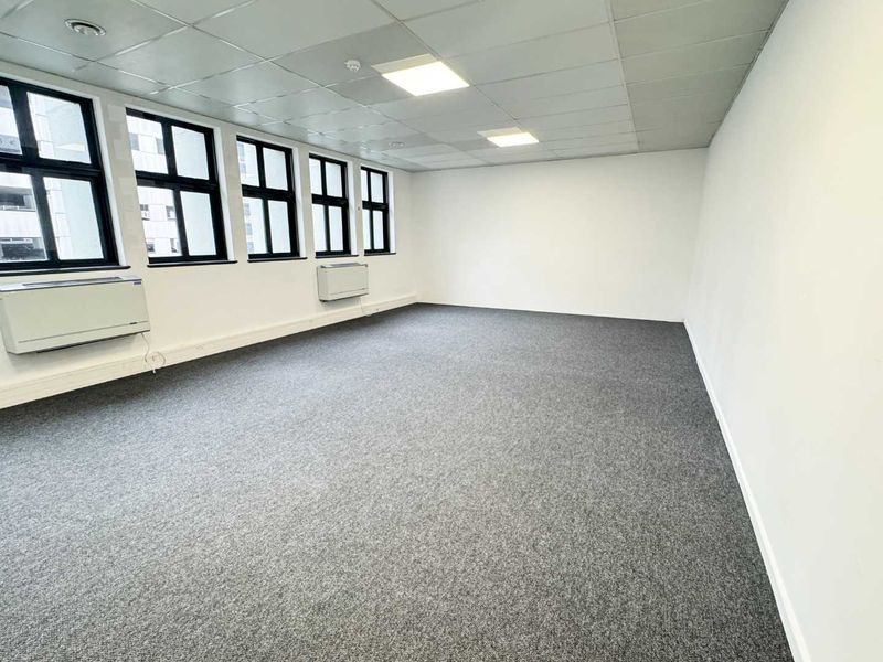 A bright, open-plan office space featuring neutral carpeting, a suspended ceiling with recessed lighting, and large windows providing ample natural light.