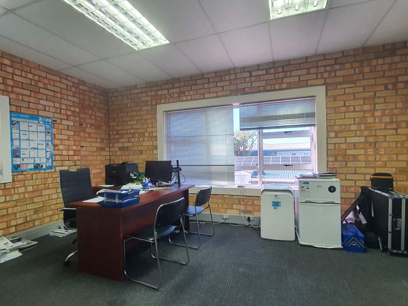 A functional office space featuring exposed brick walls, carpeted flooring, and large windows with venetian blinds, providing natural light and a professional work environment.