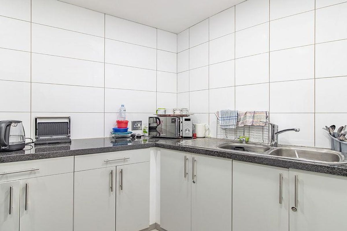 A functional office kitchenette featuring light grey cabinetry, dark granite-style countertops, and a stainless steel double sink with tiled splashbacks.