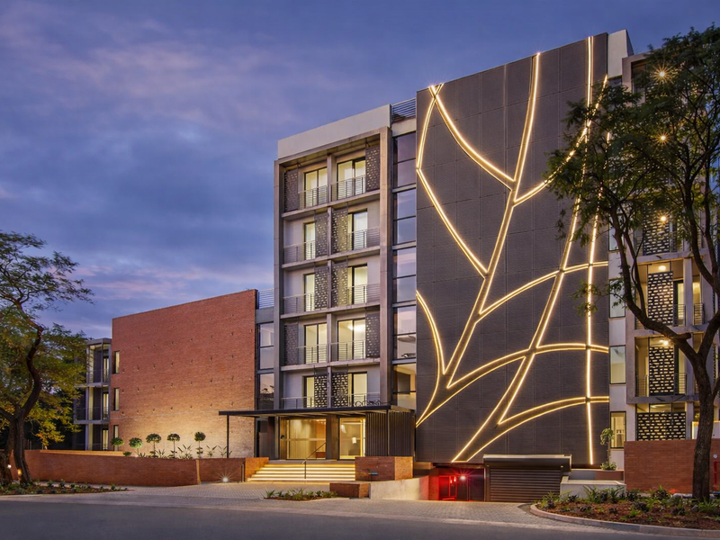 Modern apartment building exterior featuring striking architectural facade lighting, decorative metal cladding, and a secure underground parking entrance at dusk.