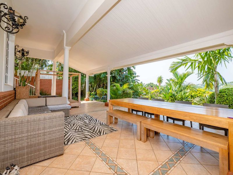 Spacious covered patio featuring tiled flooring and support columns, offering an ideal outdoor entertainment area overlooking the lush garden.