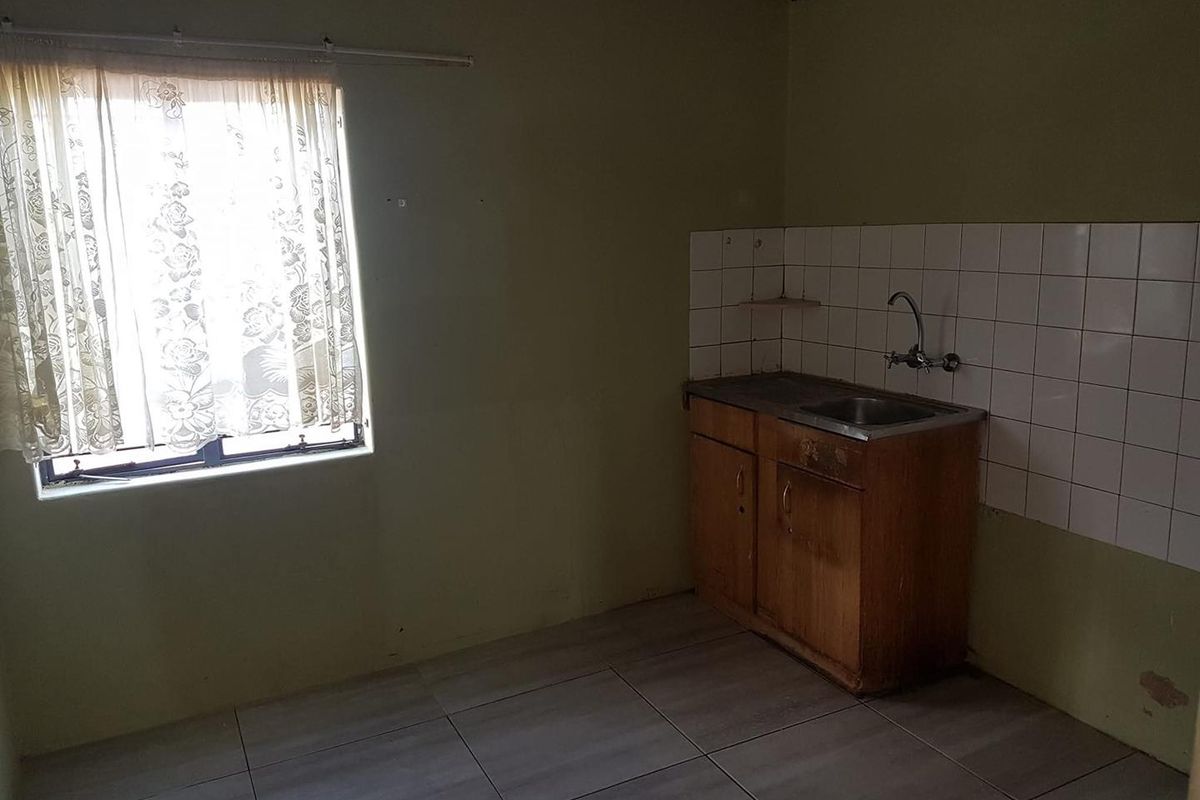 A basic, empty kitchen area featuring tiled flooring, a simple sink unit with tiled splashback, and dated cabinetry in need of refurbishment.