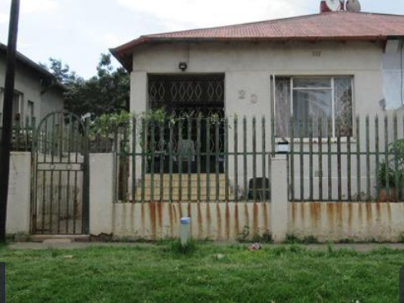 Front elevation of a single-story house featuring a covered porch, metal security fencing, and security bars on windows, showing signs of exterior wear.