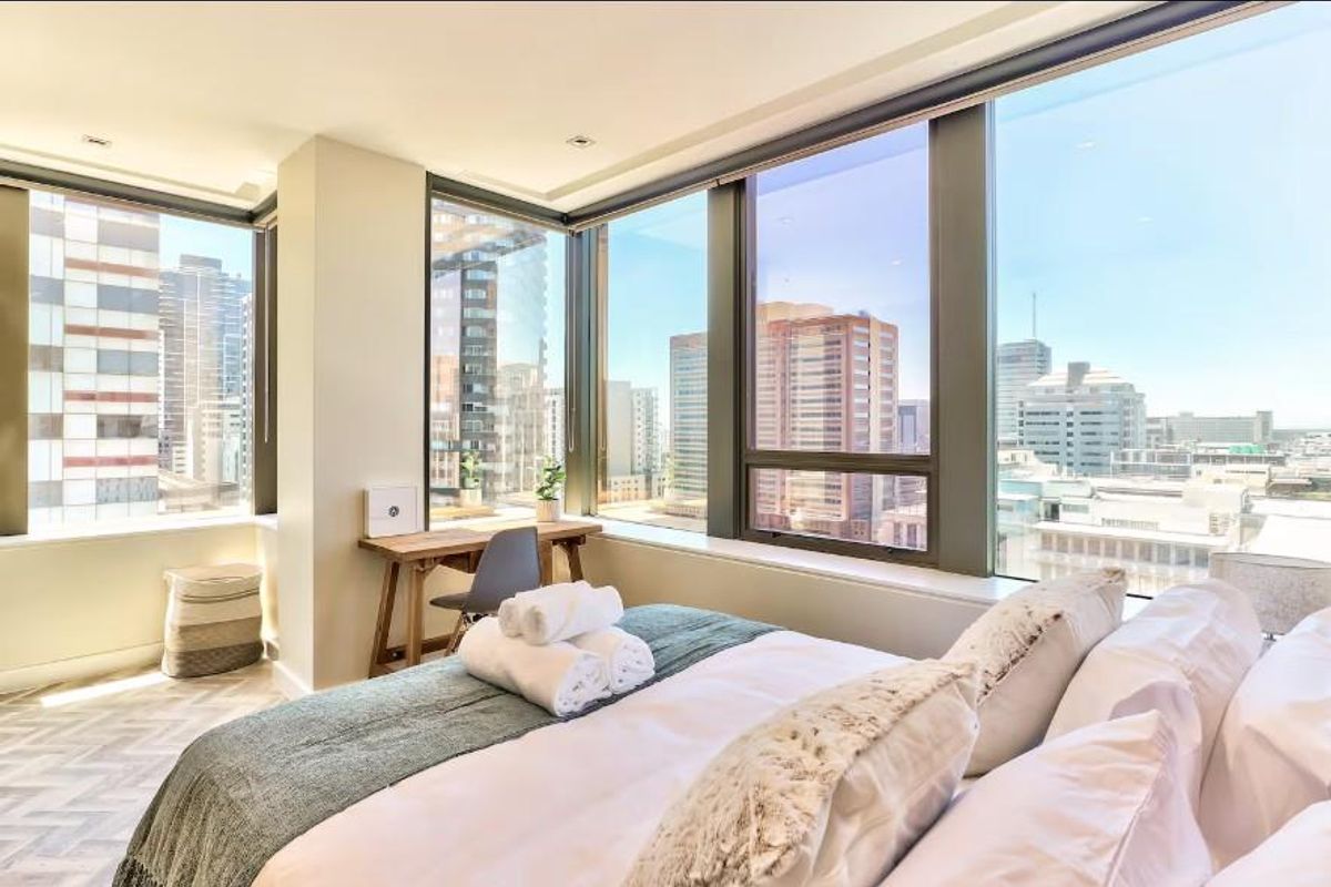 A bright, modern bedroom featuring floor-to-ceiling windows with expansive city views, recessed lighting, and high-quality flooring.