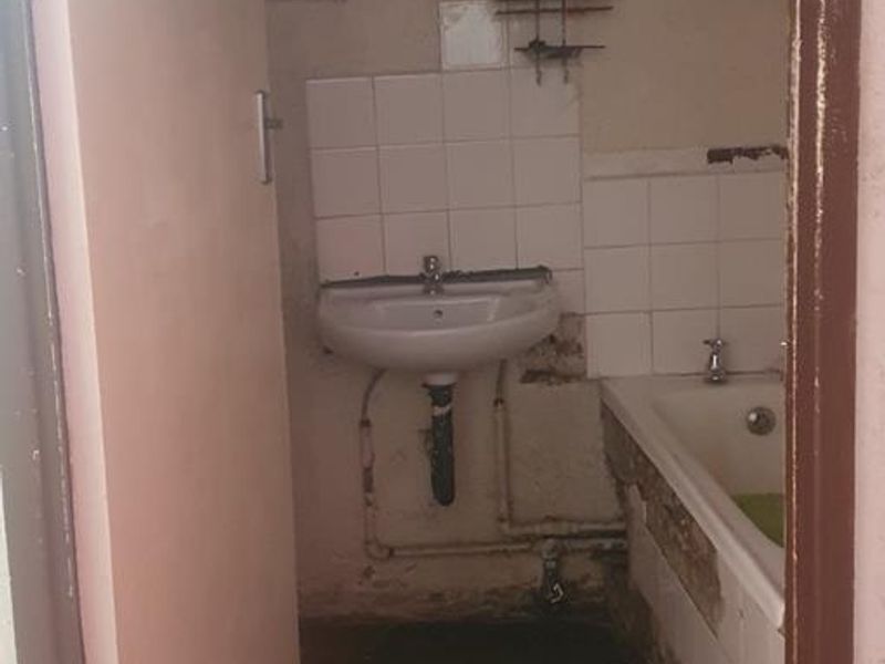 A small, dated bathroom requiring significant renovation, featuring a pedestal basin, bathtub, and exposed plumbing with visible wall damage.
