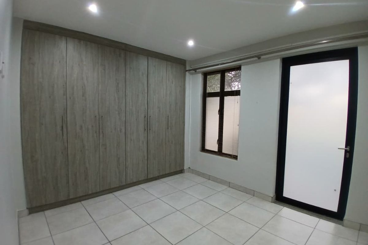 A bright bedroom featuring neutral floor tiles, built-in wooden cabinetry, recessed ceiling lighting, and a glass-panelled door leading to an outdoor area.
