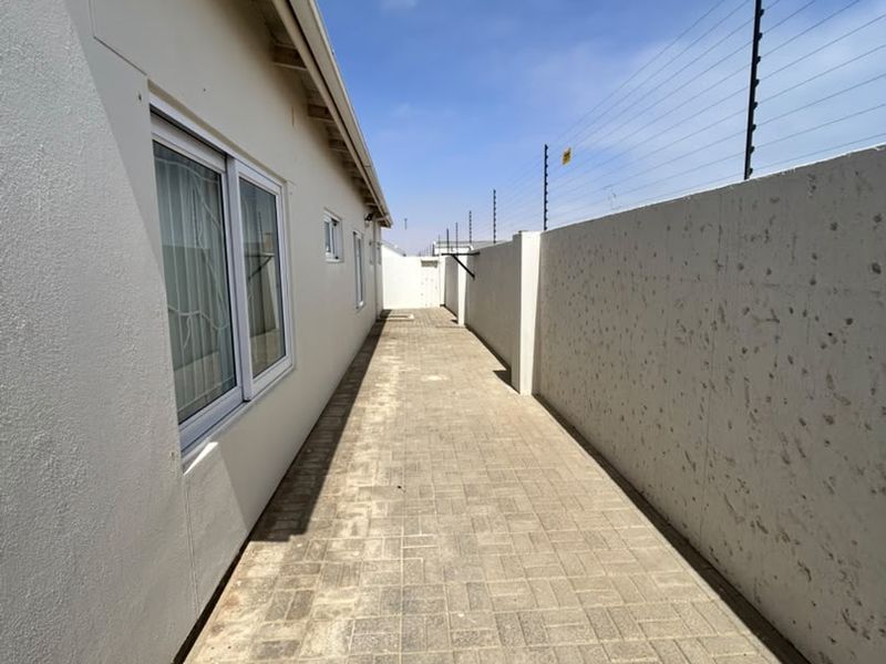 A side exterior walkway featuring paved flooring, a high boundary wall with electric fencing, and access to the home via sliding windows.