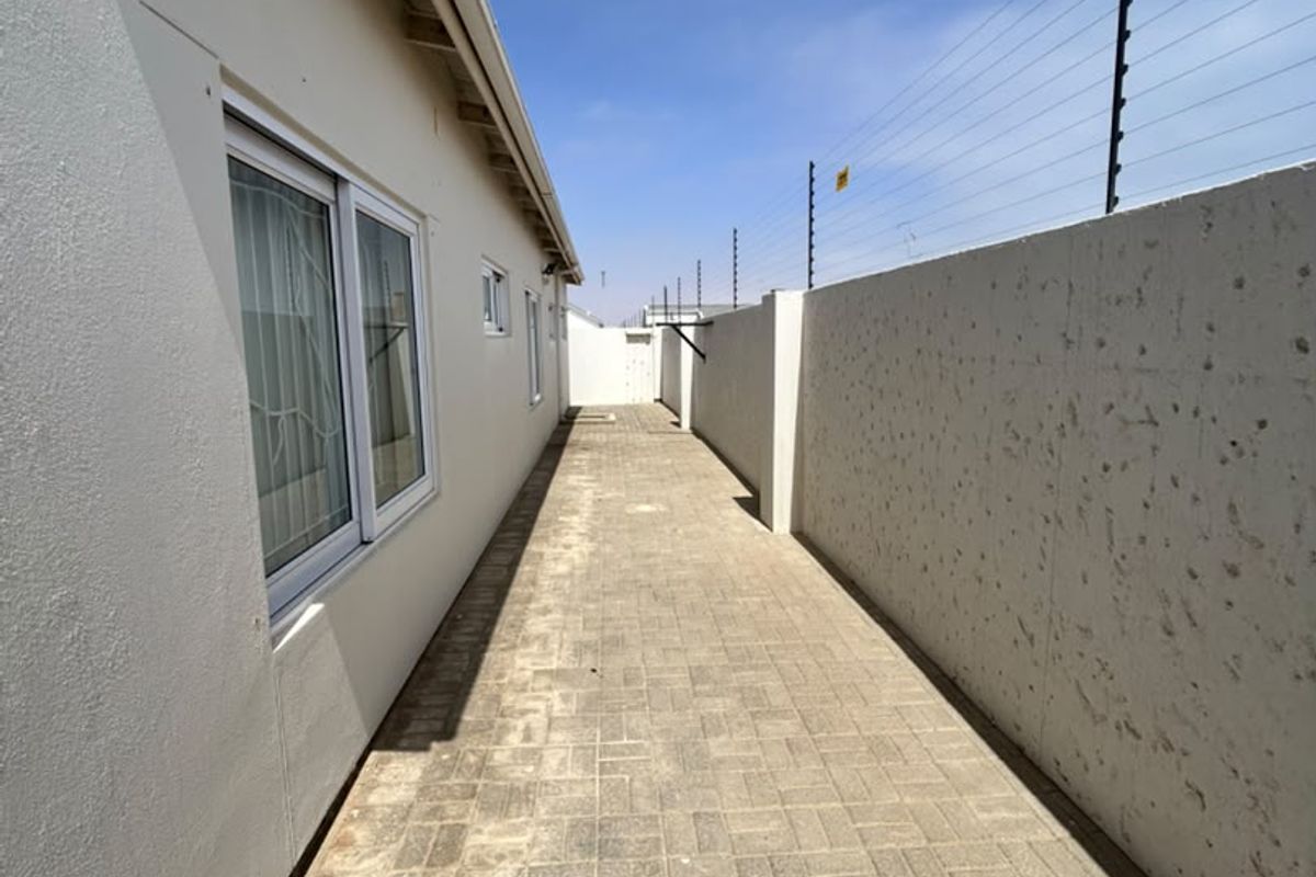 A side exterior walkway featuring paved flooring, a high boundary wall with electric fencing, and access to the home via sliding windows.