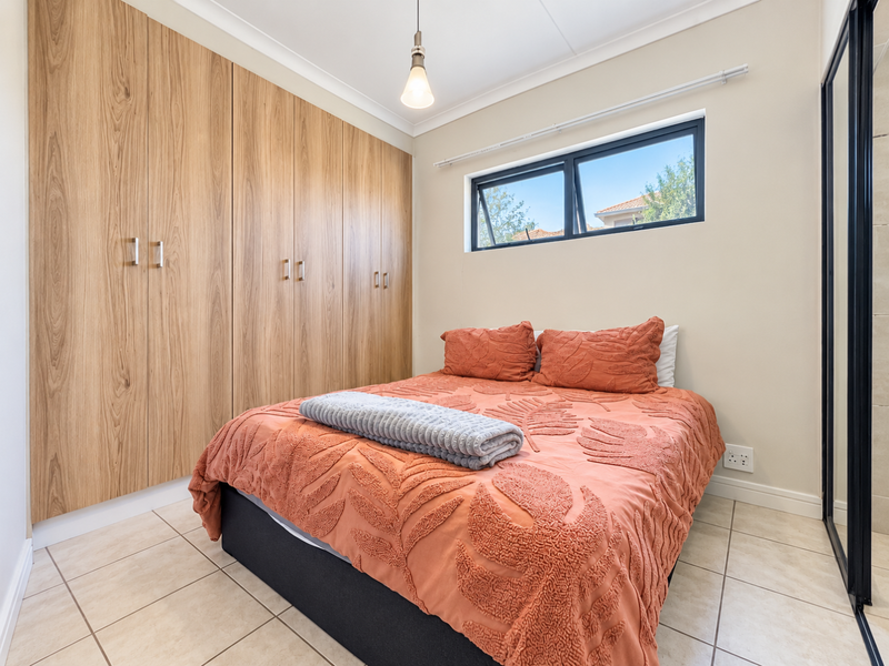 A well-maintained bedroom featuring extensive built-in wooden cupboards, neutral tiled flooring, and a top-hung window providing natural light.