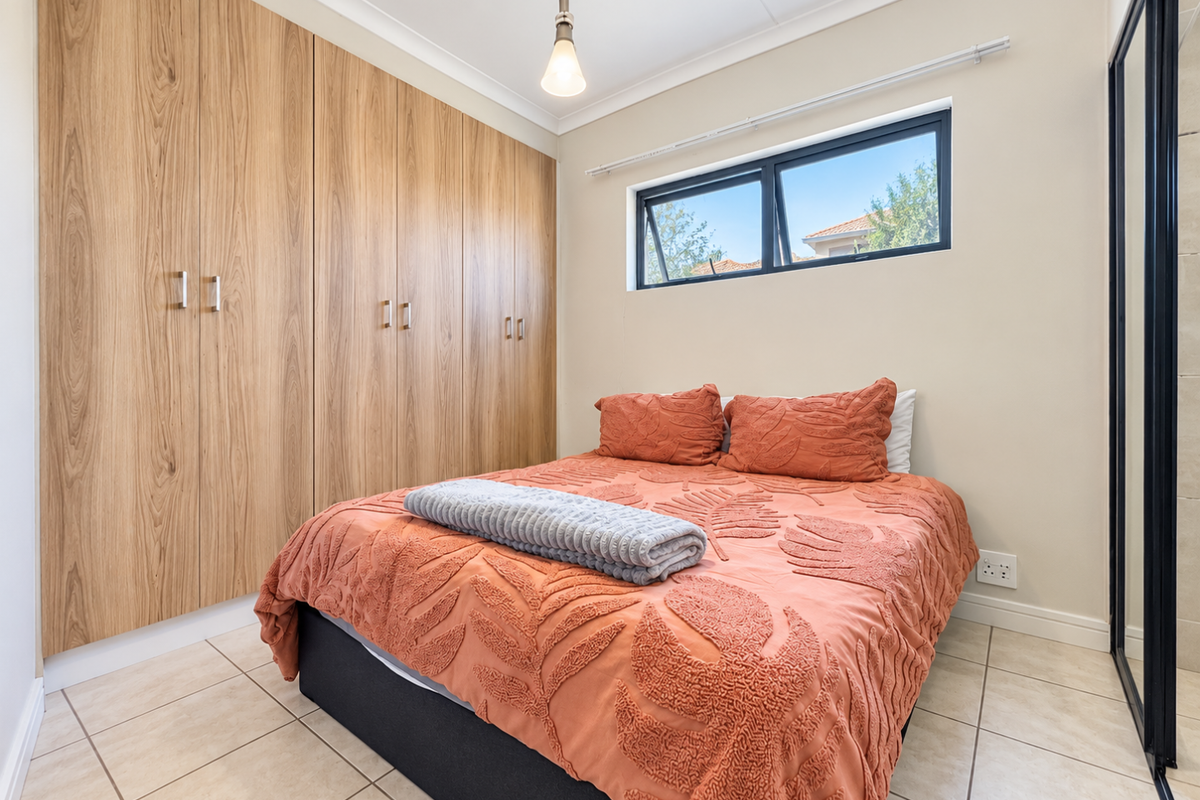 A well-maintained bedroom featuring extensive built-in wooden cupboards, neutral tiled flooring, and a top-hung window providing natural light.
