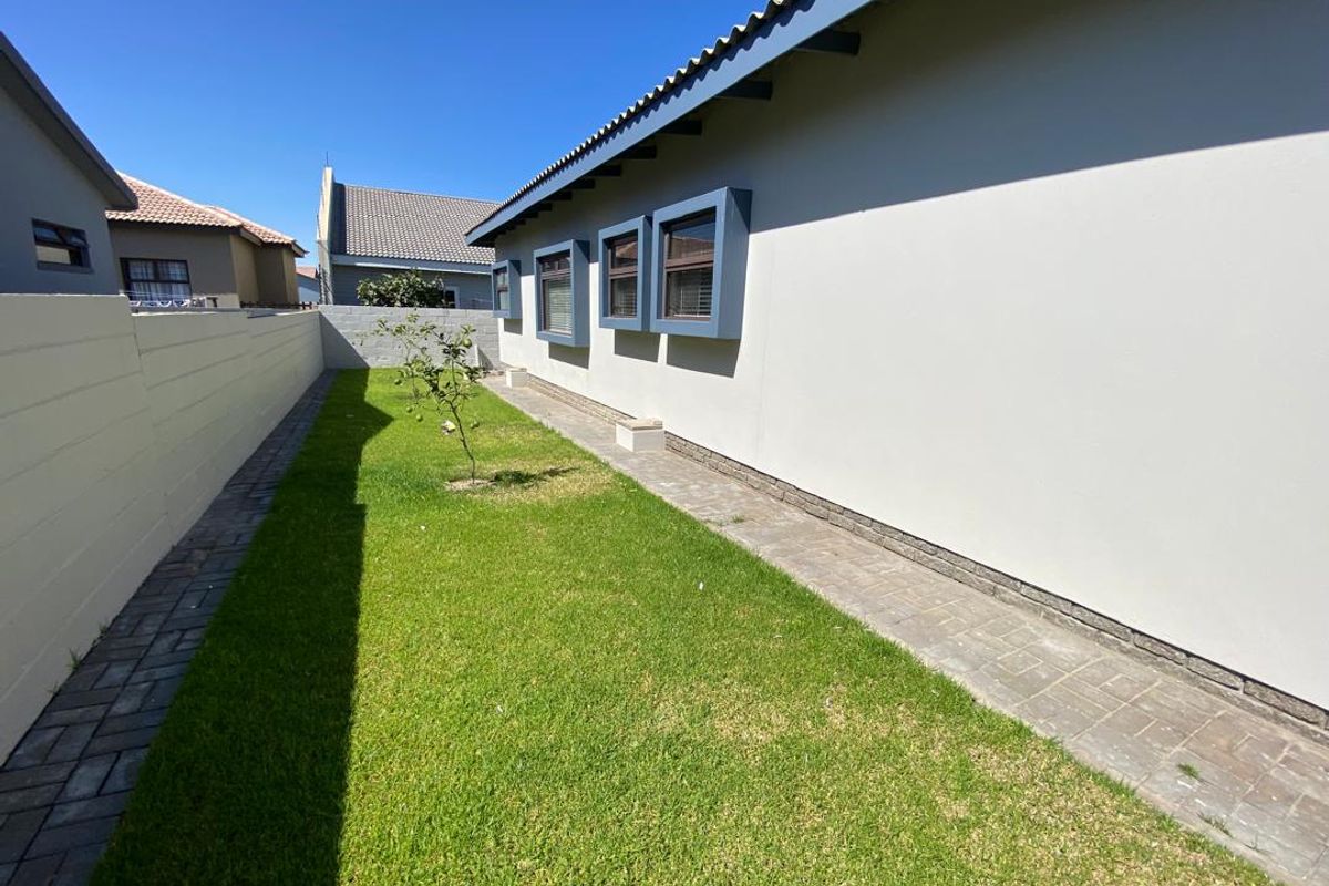A well-maintained side garden featuring a paved walkway, manicured lawn, and modern exterior wall finishes with framed window surrounds.