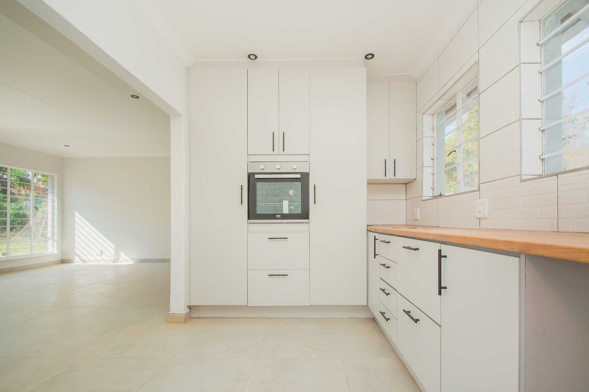 A bright, modern kitchen featuring white cabinetry, a built-in oven, and light-toned countertops, seamlessly flowing into an adjacent open-plan living area.