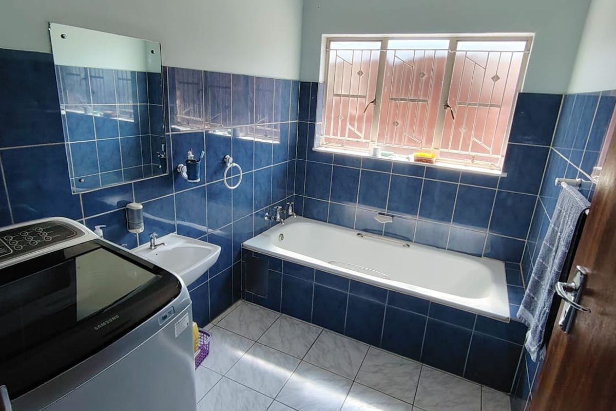 A functional bathroom featuring blue wall tiles, a built-in bathtub, a wall-mounted basin, and a window with security bars.