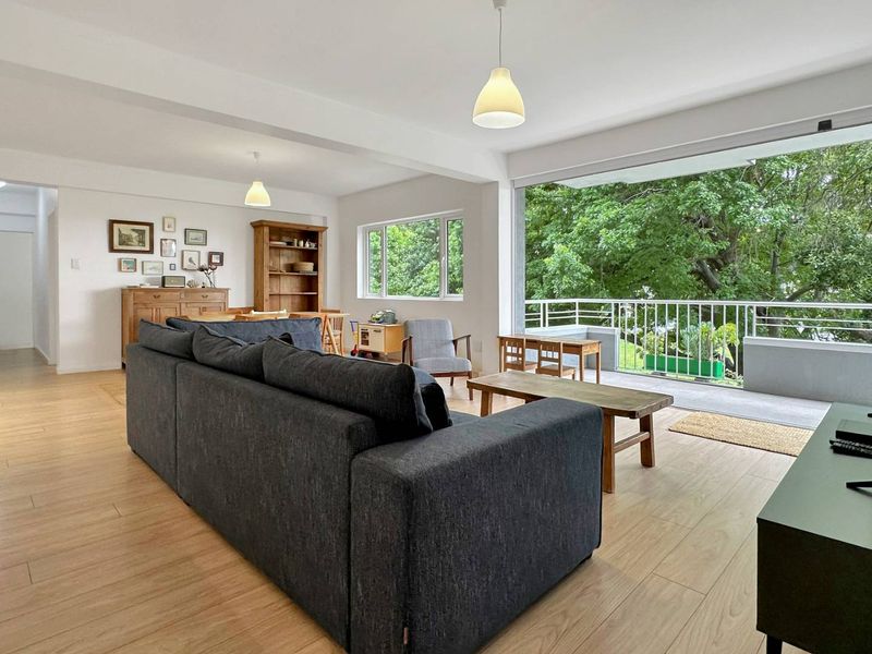 Spacious open-plan living area featuring light laminate flooring, large windows, and seamless access to a private balcony with lush green views.
